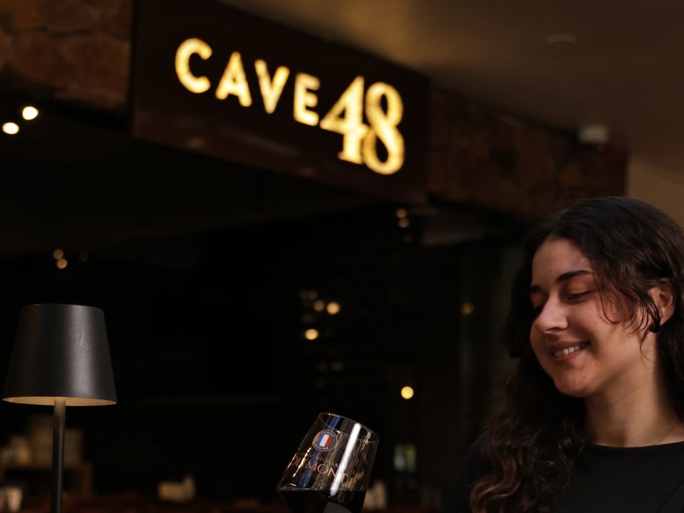 Cliente brinda com uma taça de vinho Jolimont diante do fondue, sob o letreiro do Cave 48
